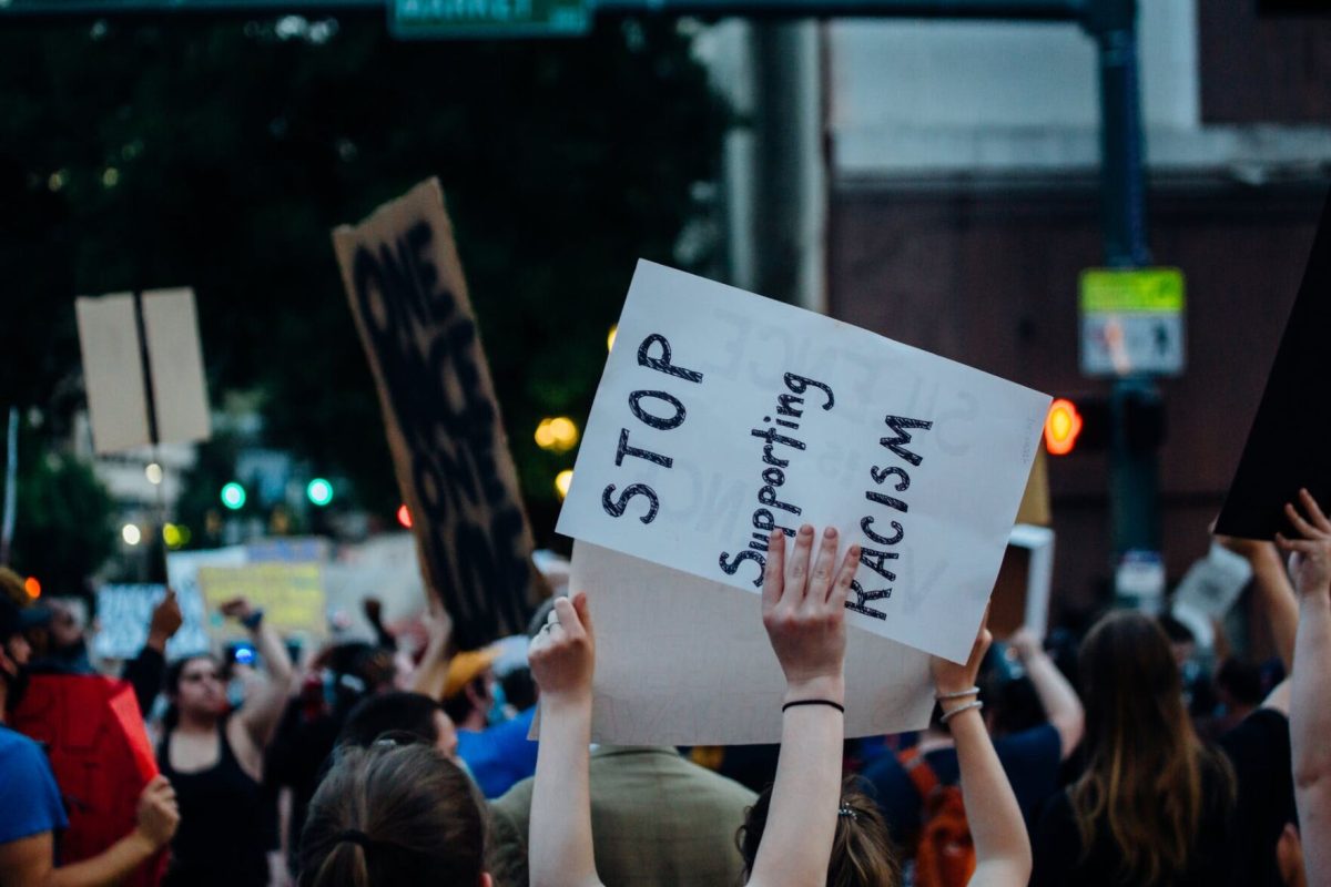 A sign with "Stop Supporting Racism" in black writing is held above a crowd.&#160;